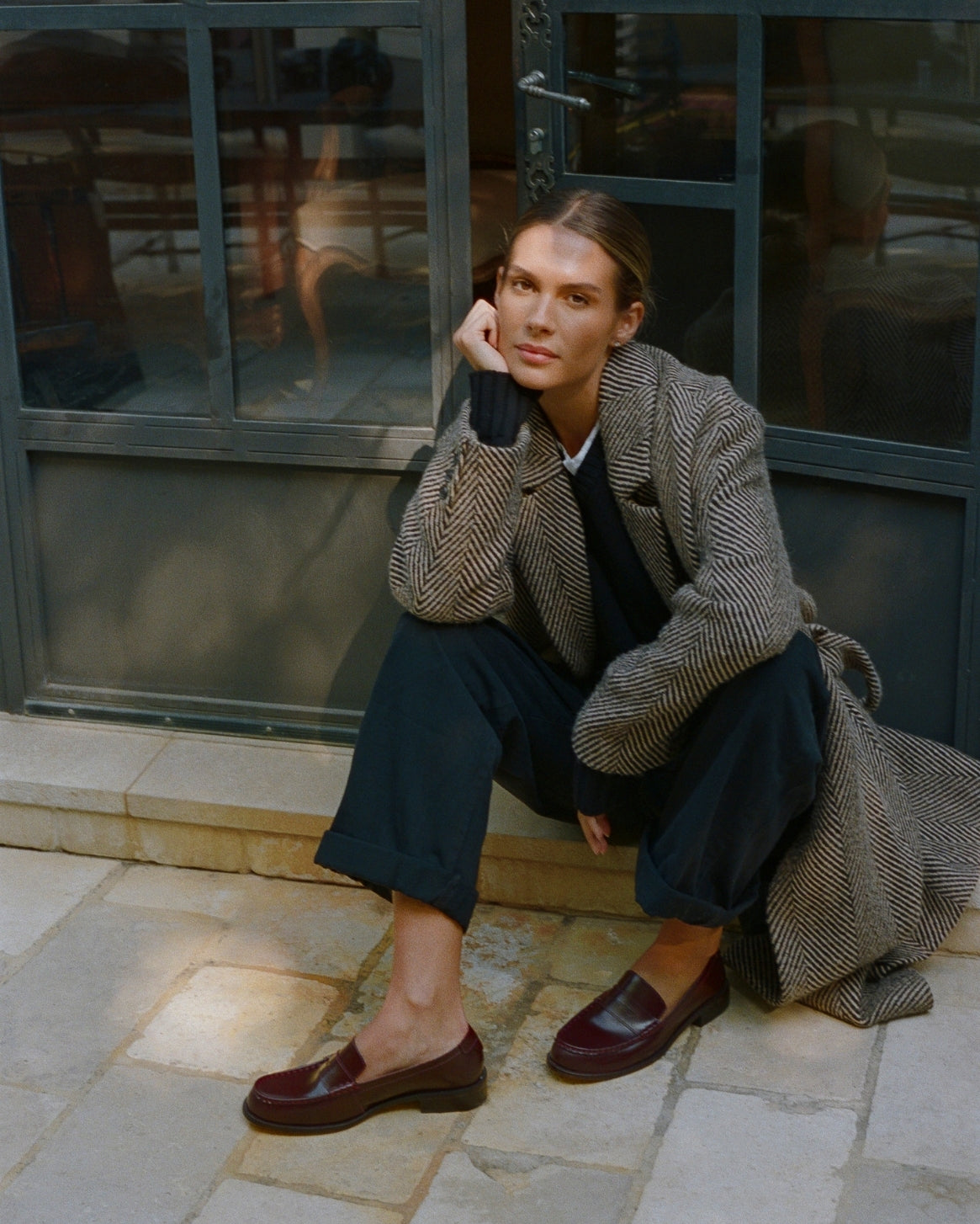 A woman crouches on stone pavement in front of a dark-framed glass door, wearing a textured gray herringbone blazer, dark navy wide-leg trousers, and dark burgundy leather loafers.