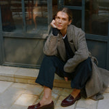 A woman crouches on stone pavement in front of a dark-framed glass door, wearing a textured gray herringbone blazer, dark navy wide-leg trousers, and dark burgundy leather loafers.
