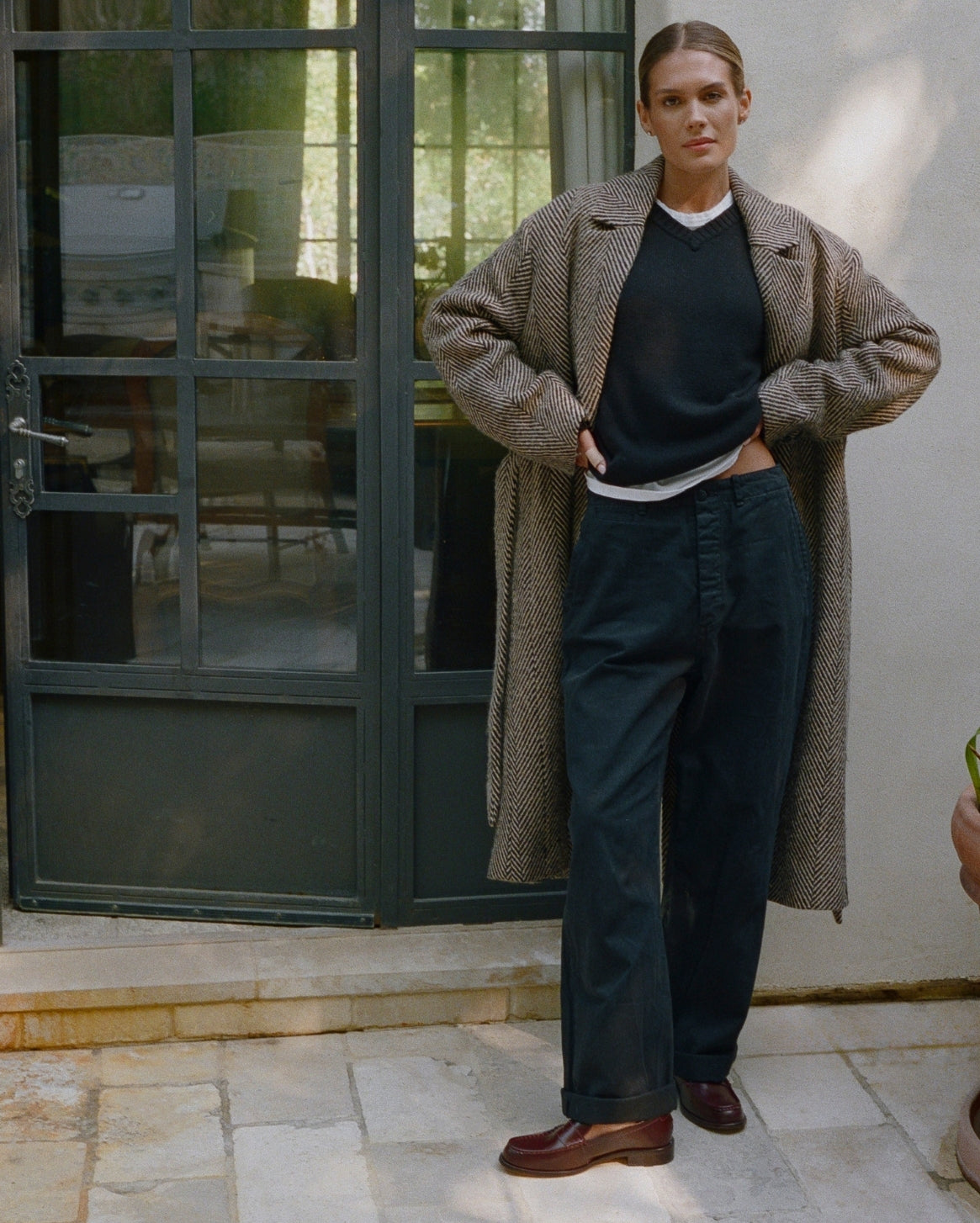 A model stands outdoors in front of a dark green glass door and light-colored wall, wearing a long beige herringbone coat over a black sweater and dark navy trousers, paired with brown leather shoes.