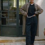 A model stands outdoors in front of a dark green glass door and light-colored wall, wearing a long beige herringbone coat over a black sweater and dark navy trousers, paired with brown leather shoes.