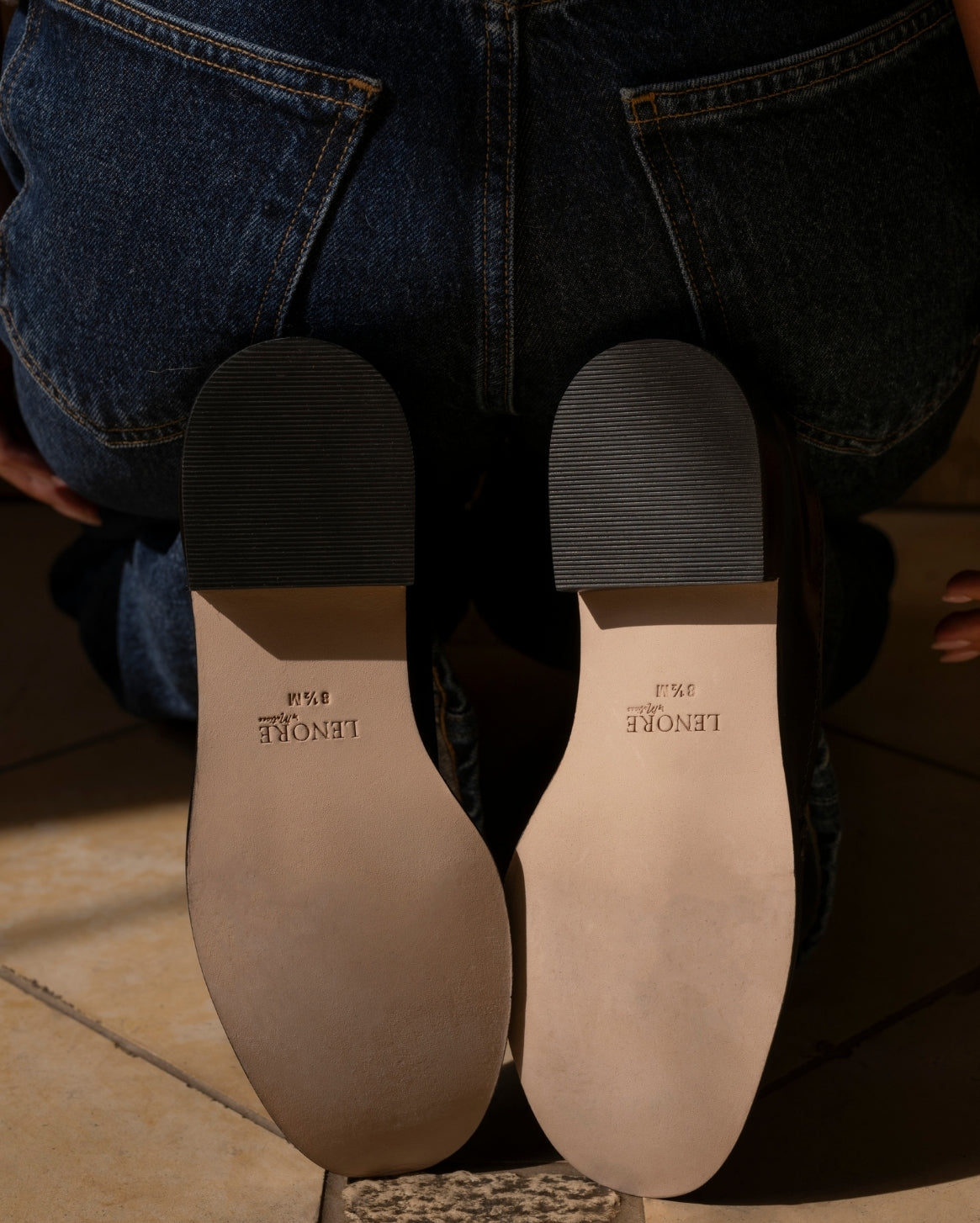 Close-up of a person wearing blue jeans with the sole of the shoes showing the brand 'Lenore'.