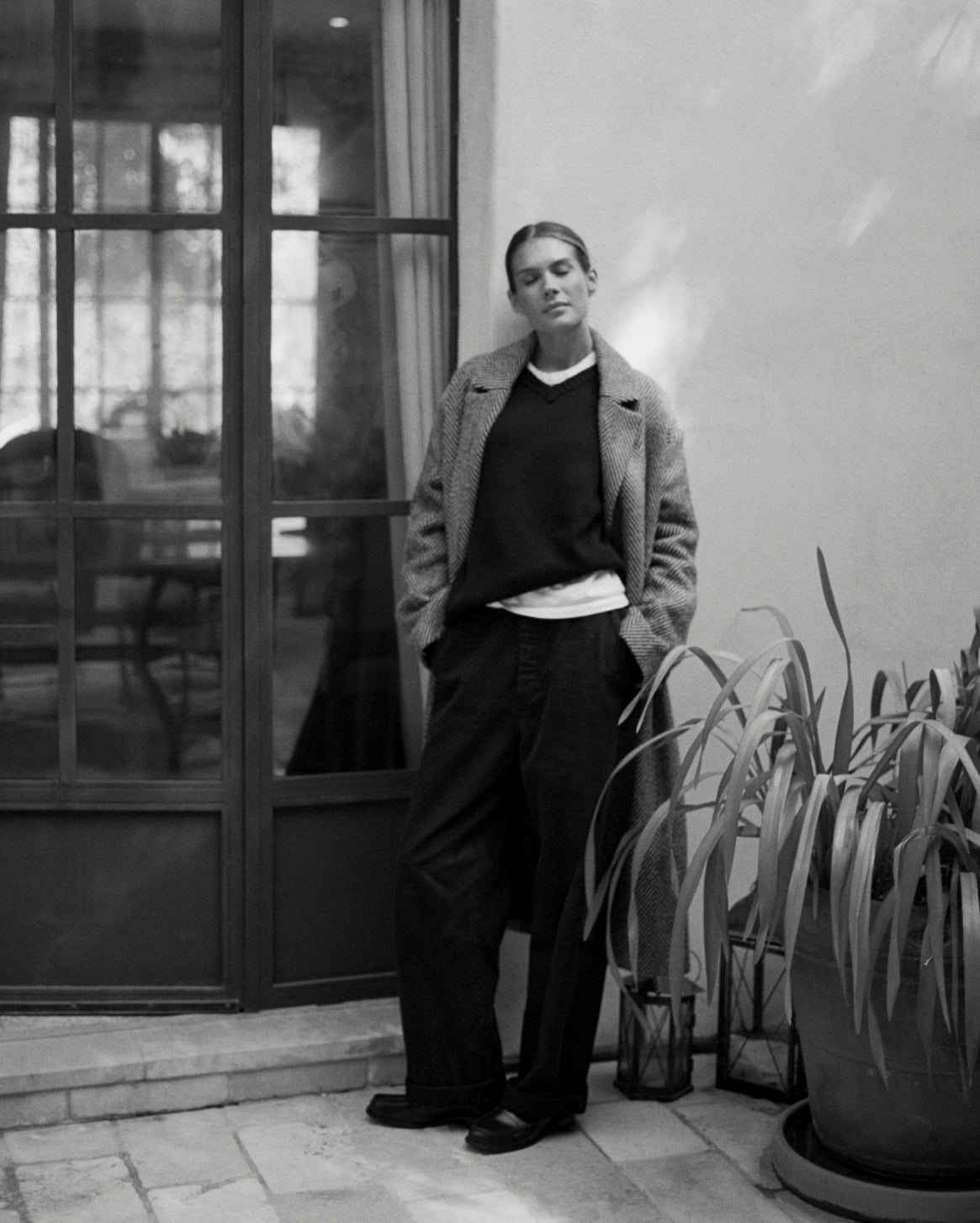 A black and white editorial photograph of a model wearing a textured tweed jacket, dark sweater, white undershirt, and wide-leg trousers, standing outdoors next to a glass door and potted plants.