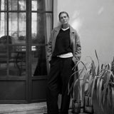 A black and white editorial photograph of a model wearing a textured tweed jacket, dark sweater, white undershirt, and wide-leg trousers, standing outdoors next to a glass door and potted plants.