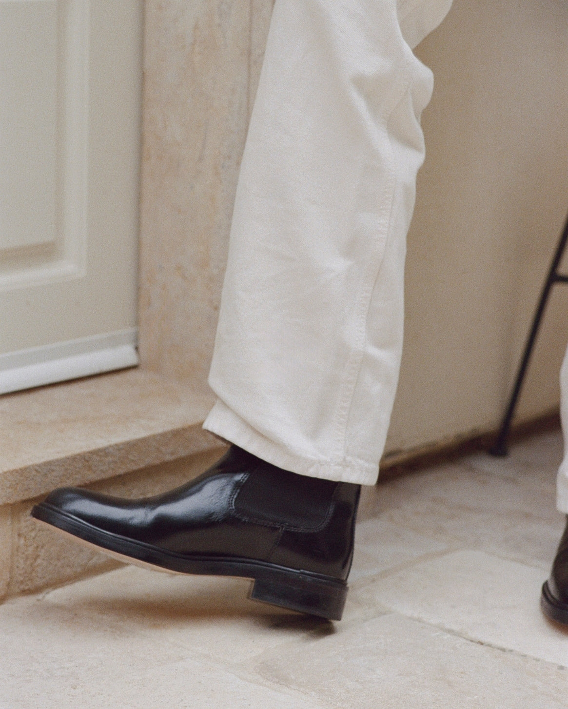 A close-up of a person's lower legs wearing off-white trousers and shiny black Chelsea boots on a stone step.