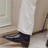 A close-up of a person's lower legs wearing off-white trousers and shiny black Chelsea boots on a stone step.