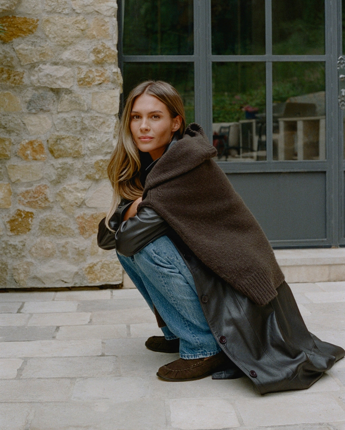 A woman crouches outdoors in front of a stone wall and large window, wearing a dark brown fuzzy hooded wrap draped over her shoulders, a black leather coat, blue jeans, and brown suede mules.