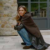 A woman crouches outdoors in front of a stone wall and large window, wearing a dark brown fuzzy hooded wrap draped over her shoulders, a black leather coat, blue jeans, and brown suede mules.