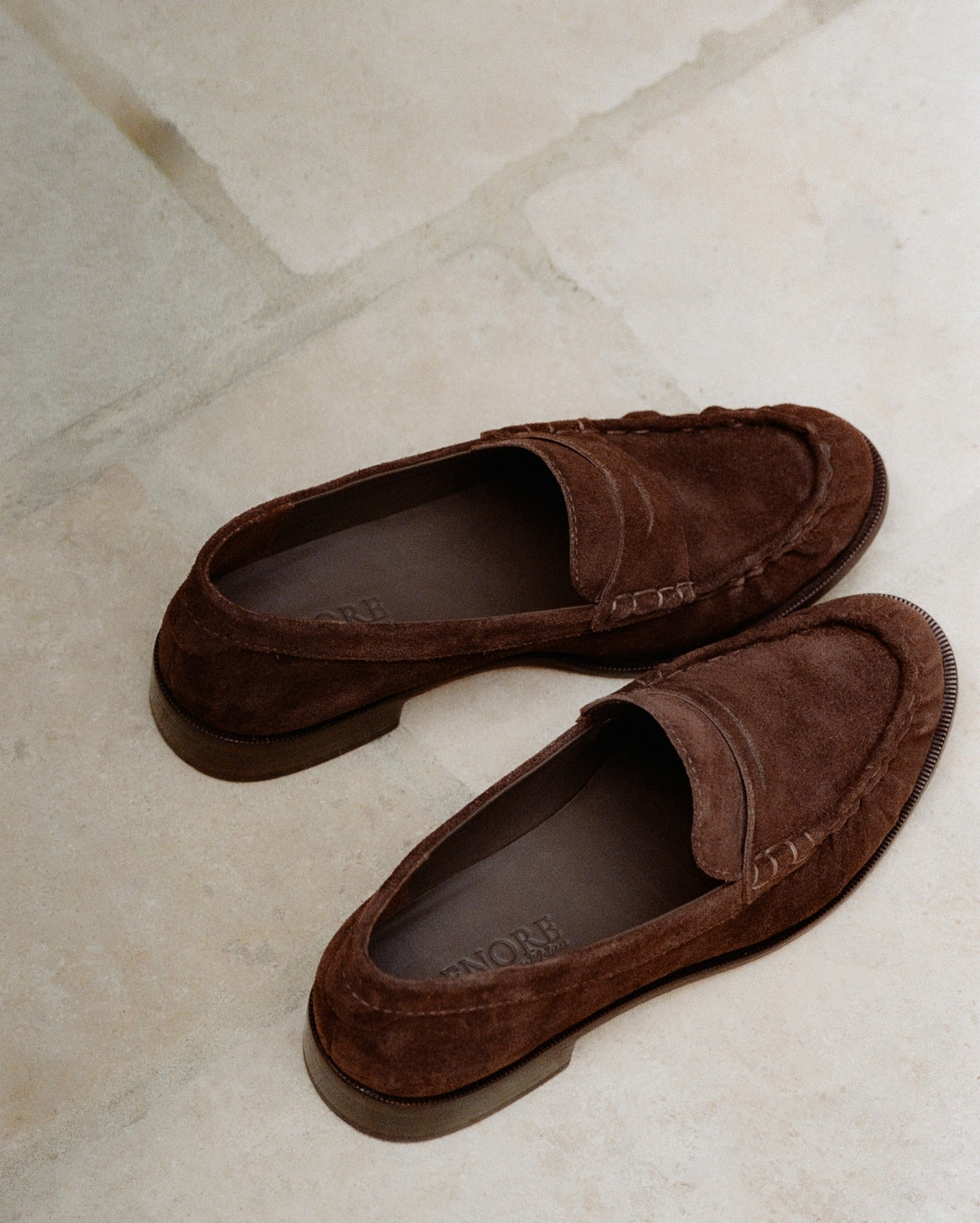 A pair of dark brown suede moccasin-style loafers photographed on a light beige stone-tile floor. The shoes are shown from an overhead angle, with one shoe positioned above the other.