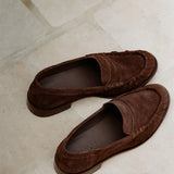 A pair of dark brown suede moccasin-style loafers photographed on a light beige stone-tile floor. The shoes are shown from an overhead angle, with one shoe positioned above the other.