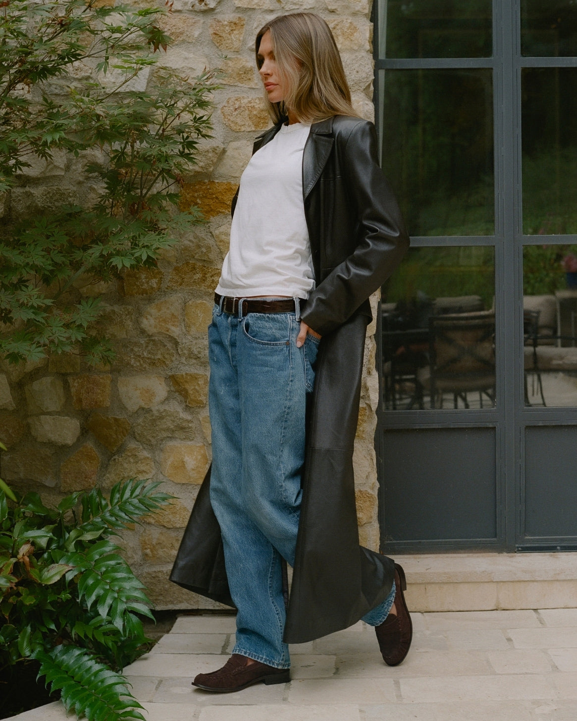 A woman stands outdoors in front of a stone wall and dark-framed glass door, wearing a long black leather coat over a white top, loose blue jeans, and brown shoes.