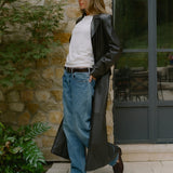 A woman stands outdoors in front of a stone wall and dark-framed glass door, wearing a long black leather coat over a white top, loose blue jeans, and brown shoes.