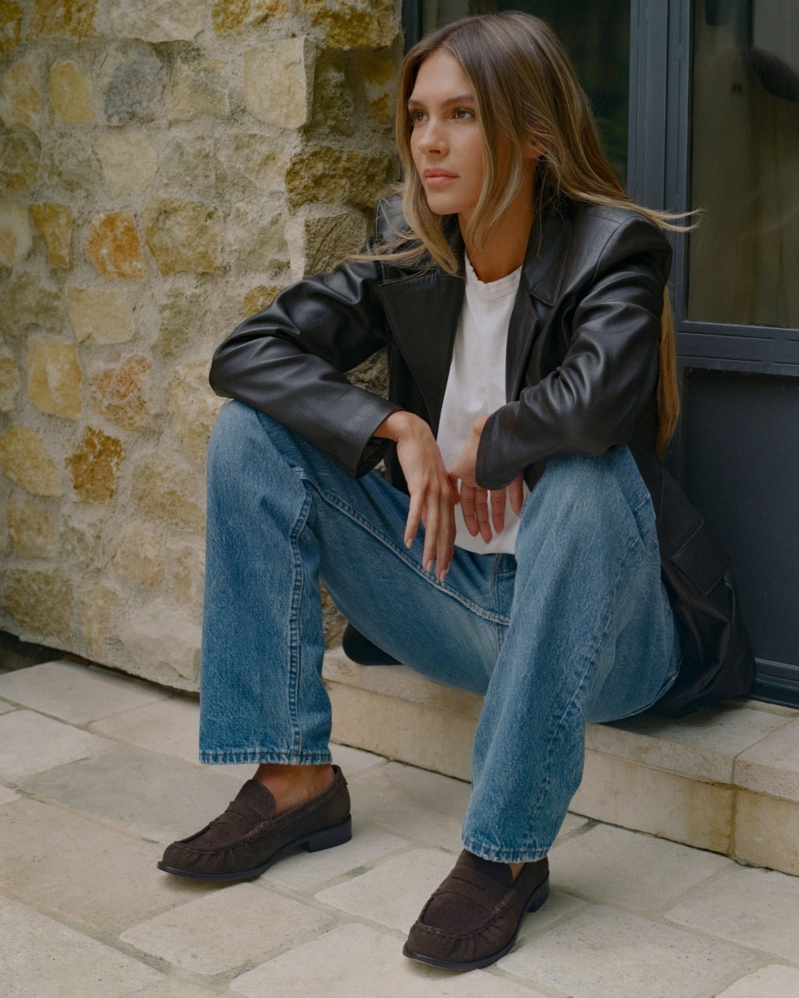A woman sits on stone steps wearing a black leather blazer, white t-shirt, blue jeans, and dark brown suede loafers, positioned against a rustic stone wall.