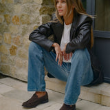 A woman sits on stone steps wearing a black leather blazer, white t-shirt, blue jeans, and dark brown suede loafers, positioned against a rustic stone wall.