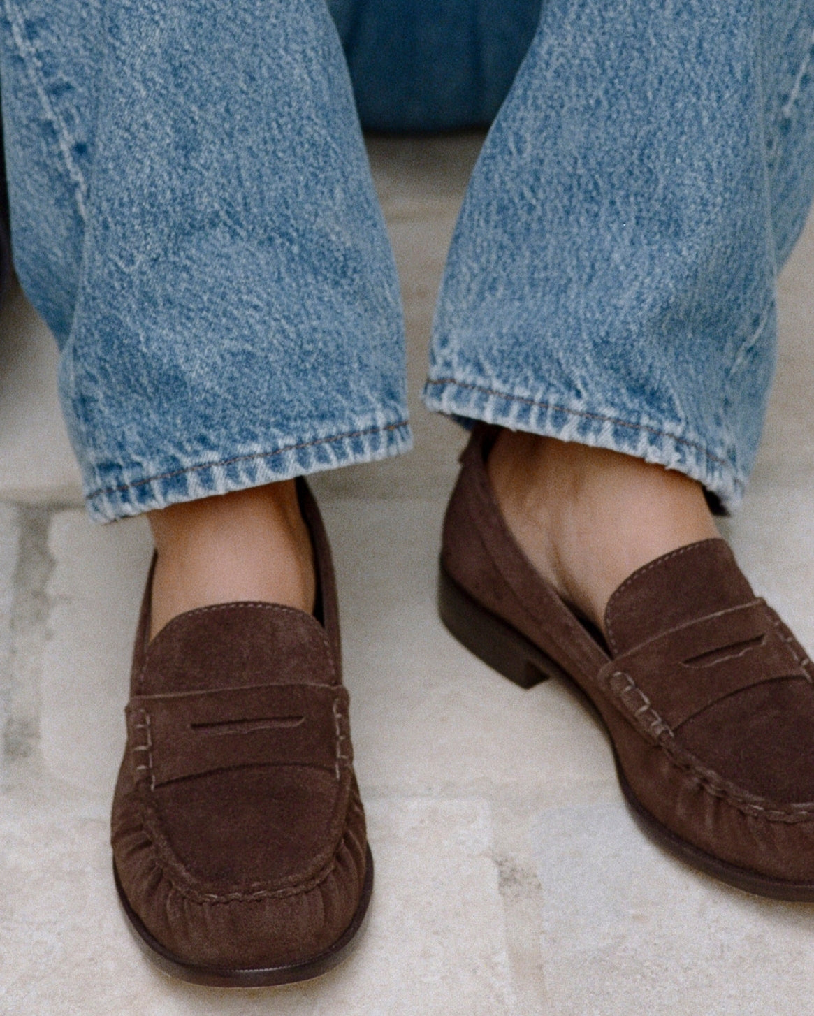 A close-up of a person wearing brown suede penny loafers with light blue denim jeans, sitting on a light stone floor.