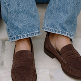 A close-up of a person wearing brown suede penny loafers with light blue denim jeans, sitting on a light stone floor.